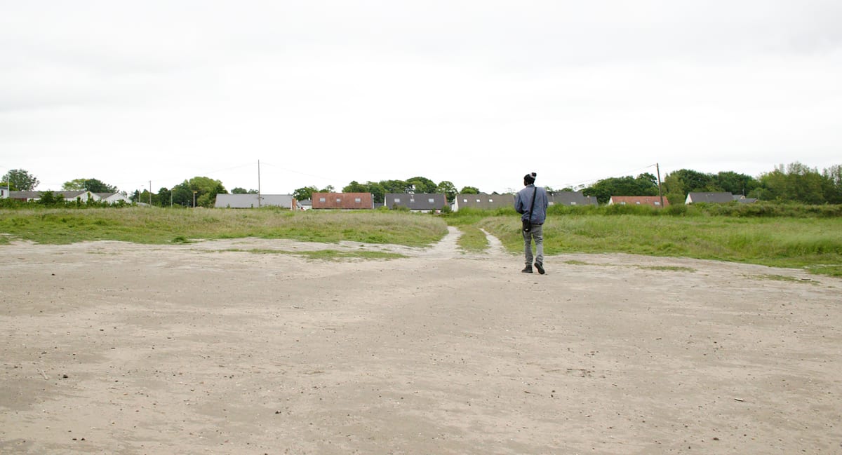 A man walks alone across open ground toward a distant village, meeting a fork in the path — a still from Nowhere To Go.