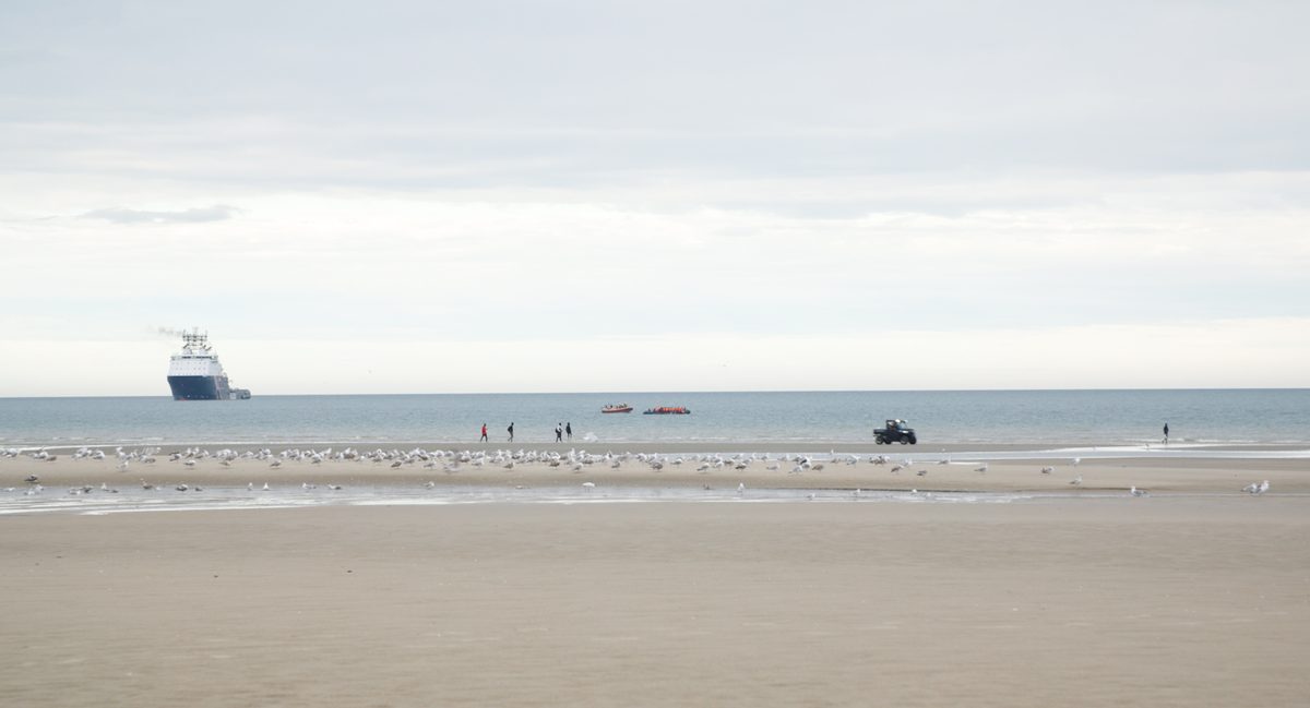 A small crowded boat is escorted off a wide beach past a flock of gulls, with a ship on the horizon. A still from Nowhere To Go.