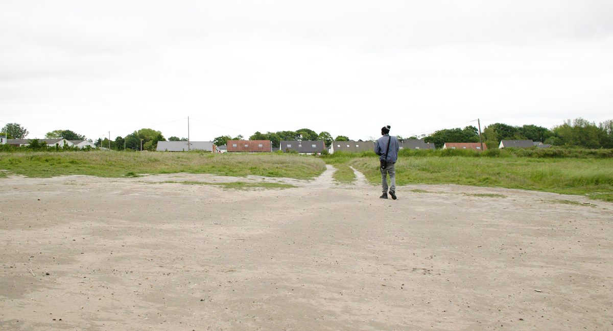 A man walks alone across open, cleared ground toward houses on the horizon, a bag over his shoulder. A still from Nowhere To Go.